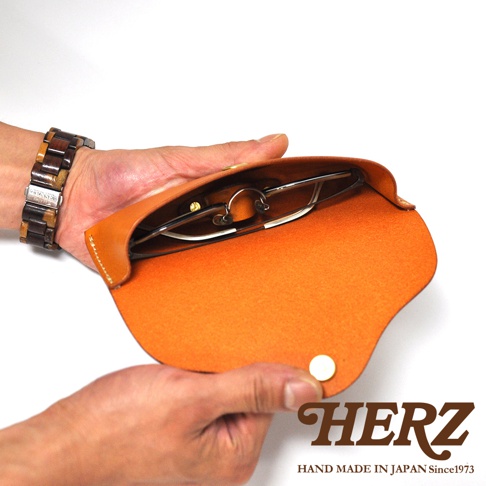 一只手拿著一副眼鏡，放入一個橘色皮革製的眼鏡收納盒。收納盒下方印有HERZ字樣和HAND MADE IN JAPAN Since1973。
