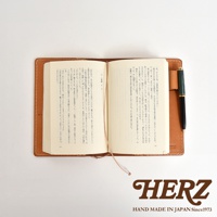 一本打開的皮革手帳，內頁有日文文字，旁放一支黑色鋼筆，手帳右下方有HERZ的Logo。