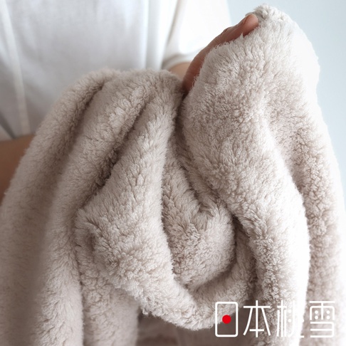 一隻手正拿著一條蓬鬆柔軟的淺駝色浴巾，浴巾有著細緻的毛圈，右下角有「日本桃雪」的紅色圓點標誌和白色文字。