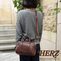 日本HERZ職人手作皮革兩用迷你波士頓包，模特背著棕色小牛皮包，包款有手提把和可拆卸肩帶，包身上印有HERZ品牌標誌。