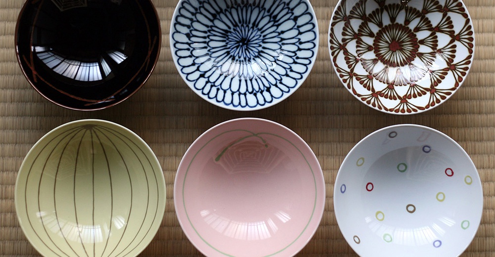 六個陶瓷平茶碗，分別為：黑色帶有紅色線條、白色帶有藍色放射狀圖案、白色帶有紅棕色花瓣狀圖案、淺黃色帶有棕色細條紋、粉紅色帶有綠色線條、以及白色帶有多色圓點。
