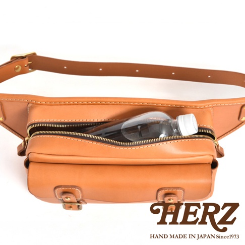 棕色皮革製作的騎士隨身斜背包，拉鍊式主袋內有一瓶透明水瓶，包身有雙層設計，搭配可調節皮帶和金屬扣環。