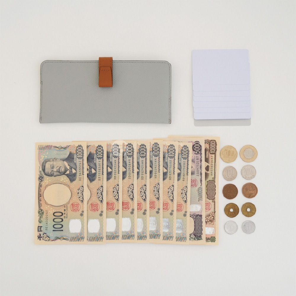 一張俯視圖展示了Atelier nuu灰色皮革雙折長夾，配有棕色皮質扣環，長夾旁整齊排列著五張空白卡片。下方平鋪八張面額一千日圓的紙鈔、一張五千日圓紙鈔與一張一萬日圓紙鈔，右側則排列著十枚不同面額的日本硬幣，呈現出極簡且收納功能完整的質感配件形象。