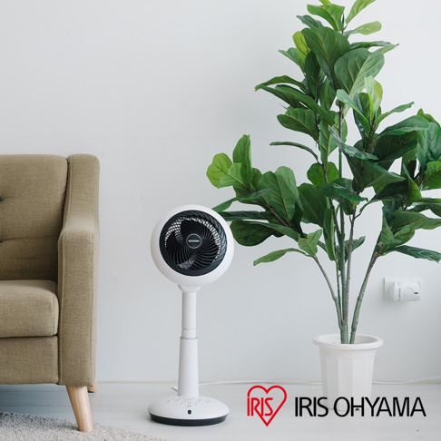白色直立式風扇，帶有黑色扇葉和黑色網罩，風扇頭可360度旋轉，底座和支架為白色。旁邊有一張米色沙發和一盆大型綠葉植物。