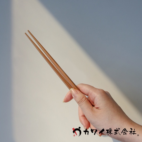 一雙約 22.5 公分的木質筷子，呈現自然的木紋與溫潤的棕色調，由手部輕巧握持，背景為淺色光滑平面，有斜向的光影。