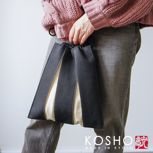 一位穿著粉色針織衫與灰色長褲的女性，手持一個京都KOSHO品牌的黑色與米白色拼接帆布信玄袋，袋身設計為獨特的摺疊風琴式結構，展現京都手作的簡約質感。