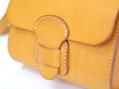 黃褐色皮革馬鞍包，帶有寬皮帶與圓形皮扣，縫線精緻，質感細膩。