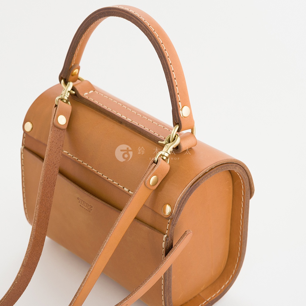 職人手作復古杜勒斯手提包，採用天然皮革製成，呈現溫潤的焦糖棕色。包身呈圓筒狀，搭配可拆卸的肩帶和提把，細節處有金屬釦和縫線裝飾，風格簡約經典。