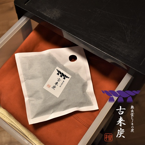 白色網狀包裝袋中的炭粒，表面印有黑色繩結圖案與「古來炭」字樣，置於橘色布料上，旁有黑色抽屜邊緣。