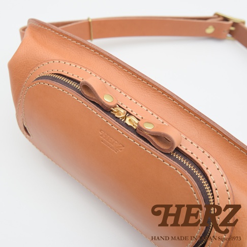 一個駝色皮革製的斜背包，有兩個拉鍊，上面有 HERZ 的品牌標誌和「HAND MADE IN JAPAN Since 1973」的字樣。