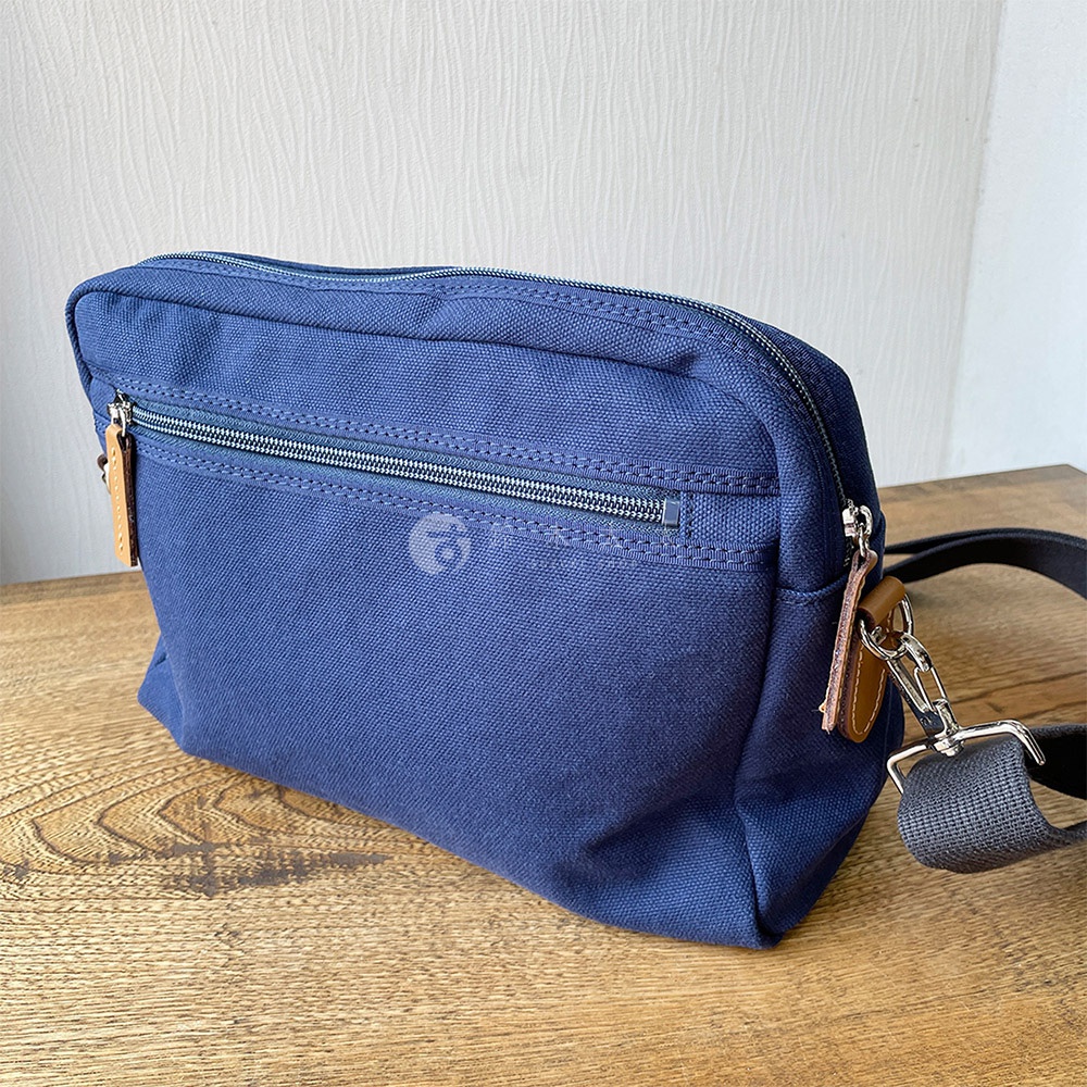 一個藍色石洗帆布側背包，正面有拉鍊口袋和一個小 logo，背包上有皮質標籤和金屬扣環。