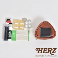 職人手作皮革小物收納包，為三角飯糰造型，主體為棕色皮革，正面縫有黑色皮革飾片，搭配金色拉鍊。收納包旁擺放有藥品、藥膏、貼布及一小盒藥丸。