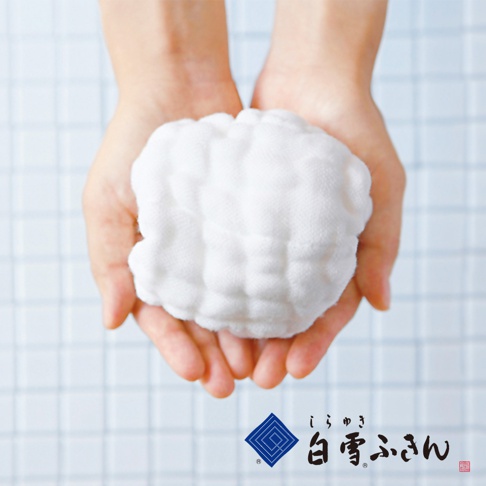 一雙手捧著白色、蓬鬆、多層次的棉質洗澡巾，背景為淺藍色格紋。
