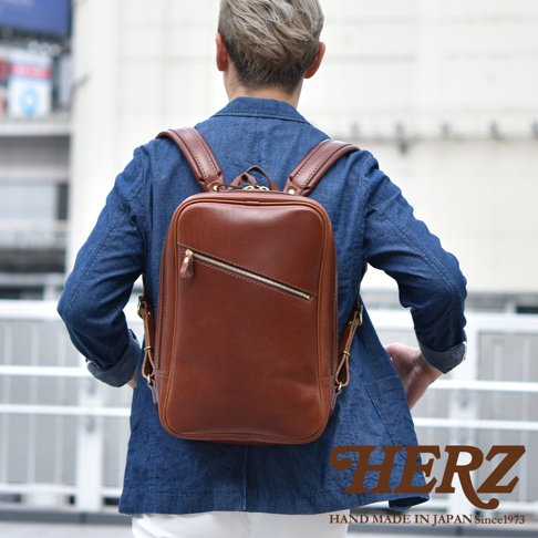 職人手作皮革長方型後背包，採用鞣製皮革材質，呈現深棕色調。包款設計簡約，正面設有斜向拉鍊口袋，肩帶與包體皆有皮革拼接與縫線細節。