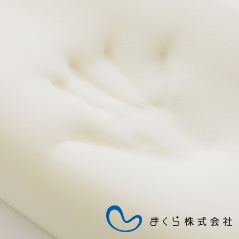 米白色記憶棉半身枕，表面有明顯的壓痕，質地柔軟。右下角有藍色曲線標誌及日文「まくら株式会社」字樣。
