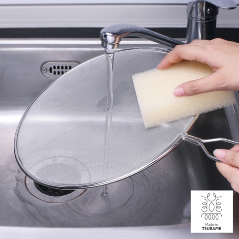 一只手正在用黃色海綿清潔一個銀色的金屬網狀透視防噴濾網，濾網置於水龍頭下方、流理臺水槽中，濾網邊緣有金屬框和手柄。