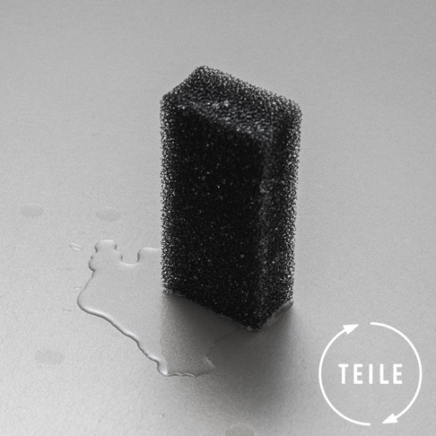 一塊黑色超細纖維海綿，具有細密的網狀紋理，放在淺灰色背景上，周圍散落著水滴。海綿底部邊緣有白色水漬。右下角有一個白色圓形箭頭標誌，其中包含文字「TEILE」。