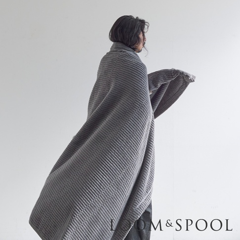 一位人士披著一件灰色的 GRAU 立體直紋純棉微厚單人毯，毯子質地柔軟，有細緻的垂直壓紋。