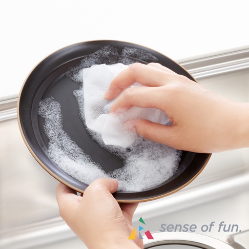 一隻手拿著白色網狀洗碗巾，正在清潔一個有泡沫的黑色盤子。另一隻手扶著盤子。