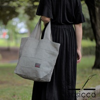 一位女性手提一個灰色帶有黑點刺子織紋路的帆布托特包，包身有水平縫線，左側有紅色品牌標籤。