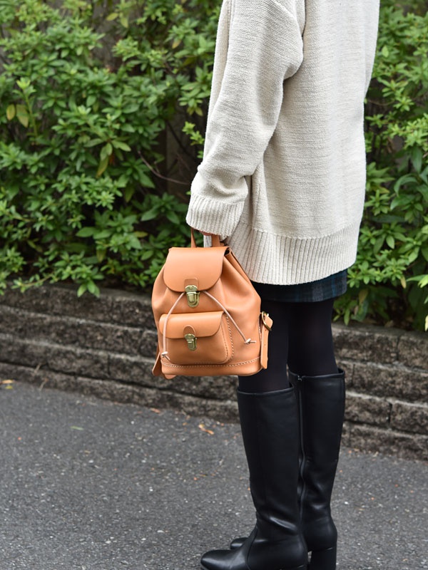 一個手持淺棕色皮革迷你後背包的人，背包上有兩個前袋和金屬搭扣，背景是綠葉和石牆。