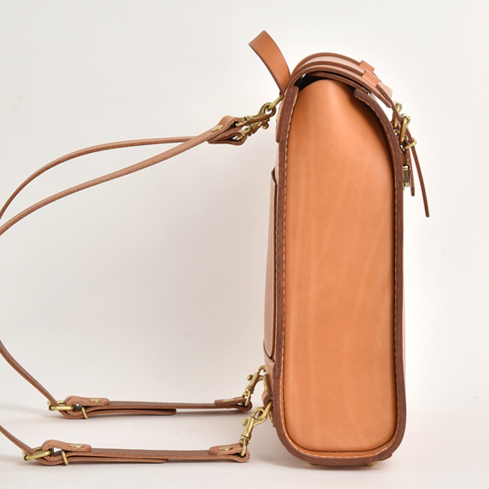 側邊視角展示一個 tan 色皮革後背包，厚實的皮革材質，帶有兩條可拆卸的皮革肩帶，肩帶與包身以黃銅色釦環連接，包身邊緣有深棕色縫線。