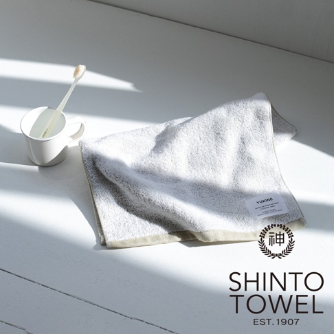 YUKINE 雪中漫步有機棉蓬厚毛巾，淺灰色，質感蓬鬆柔軟，帶有品牌標籤，旁邊有一個白色馬克杯，杯中有牙刷。