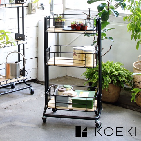 黑色金屬三層置物推車，搭配木質層板，其中一層擺放有植物、罐子和噴霧瓶，另一層有罐子，最底層有收納盒、綠色盒裝物及布料。推車底部有四個輪子。