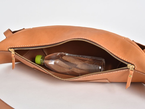 職人手作皮革斜背包，淺棕色，主隔層拉鍊打開，內部可見一瓶透明塑膠瓶裝水。包包採用車縫線工藝，材質為皮革。