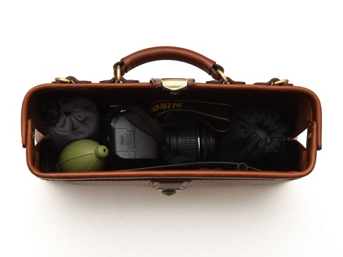 職人手作皮革橫長形杜勒斯單肩包 (CW-4)，內裝有黑色相機、綠色吹氣球、灰色和黑色收納袋，以及 Nikon 相機背帶。包身為棕色皮革材質，配有金色金屬鎖扣和提把。