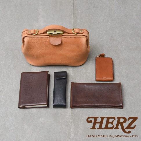 日本HERZ職人手作皮革，展示了多款棕色與黑色系的各式皮革製品，包含一個大型手提包、一個長夾、一個短夾、一個筆套及一個筆記本，呈現精緻的皮革工藝。