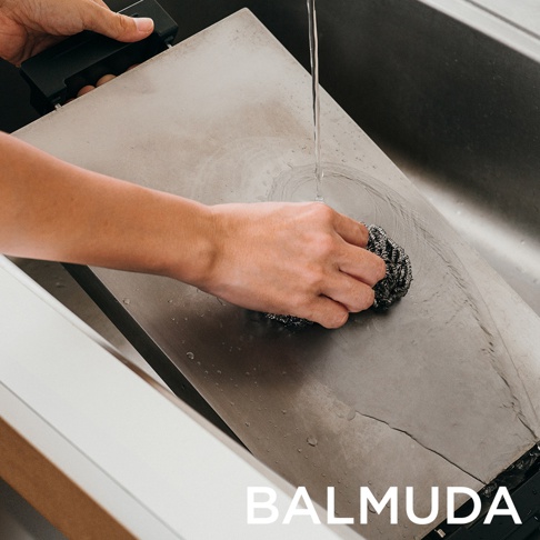 手持金屬清潔球，在水龍頭下清洗黑色極致電烤盤的金屬表面。