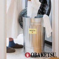 OBAKETSU 品牌附輪垃圾桶，裝有白毛巾，旁邊是洗衣機，地板上有室內拖鞋。