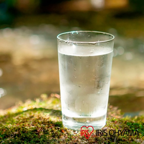 玻璃杯裝滿清澈透明的礦泉水，杯身附有水珠，放置於佈滿苔蘚的岩石上，背景為模糊的自然景觀。
