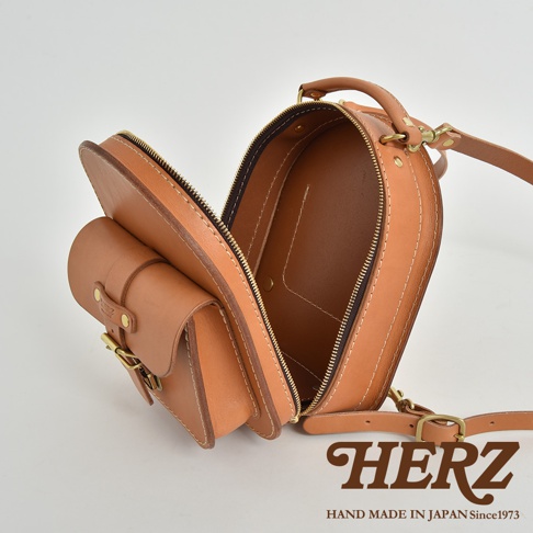 職人手作皮革迷你後背包，展示自然色澤的皮革材質，整體為圓潤的橢圓造型，設有拉鍊開口和前置小口袋，口袋處有金屬扣環。