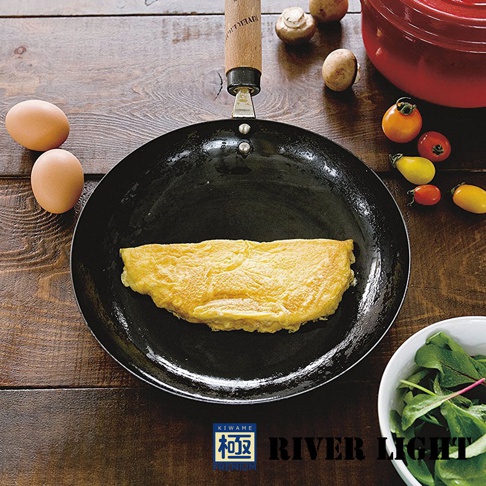 黑色平底鍋中煎著金黃色的歐姆蛋，鍋身旁有兩顆雞蛋、幾顆小番茄、蘑菇和一碗綠葉蔬菜。鍋柄為木質，鍋身有「KIWAME」和「RIVER LIGHT PREMIUM」的字樣。