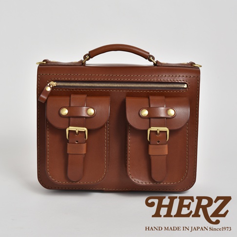 棕色皮革雙口袋兩用單肩包，正面有兩個帶有黃銅釦環的翻蓋口袋，上方為拉鍊開口，頂部有提把。