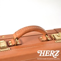 職人手作的淺棕色皮革手提行李箱，帶有黃銅色五金配件，包含提把、鎖扣和鉚釘，縫線清晰，展現英倫風格。