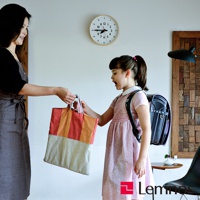 一位穿著灰色圍裙的女性將一個紅白條紋帆布袋交給一個穿著粉色碎花洋裝、背著黑色日式書包的小女孩。牆上掛著一個木製邊框的圓形時鐘。