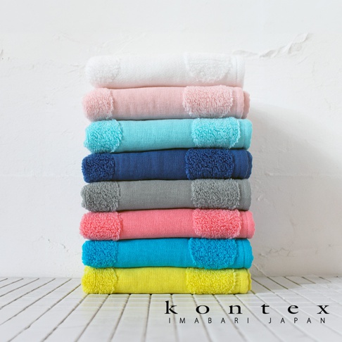 一疊色彩繽紛的 Kontex 今治毛巾垂直堆疊擺放在白色磁磚面上，背景為白色的斑駁牆面，毛巾呈現由上至下依序為白、淡粉、淺藍、深藍、灰、珊瑚紅、亮藍及亮黃的排列，底部印有 KONTEX IMABARI JAPAN 的黑色品牌字樣。