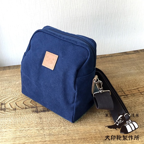 一個深藍色石洗帆布製成的方形散步包，包身上有一個淺棕色皮革材質的方形標籤，上面印有可愛的貓咪圖案和一些文字，包包配有黑色帆布肩帶，肩帶上有金屬掛鉤，背景是淺色的牆壁和木紋桌面。