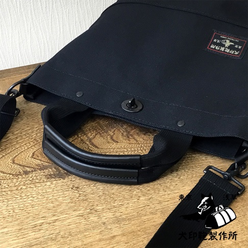 黑色帆布托特包，配有黑色皮革提把、可拆卸肩帶及旋轉鎖扣。包身印有「犬印製作所」的紅色標籤。