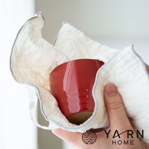 一塊米白色、帶有灰色滾邊的棉混紗廚房拭巾，包覆著一個紅色的馬克杯。拭巾表面有壓紋，質地柔軟。