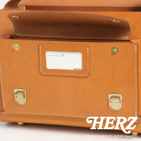 近拍皮質橫式包包，可見側邊銅質鎖扣、縫線細節，前方有一個附有標籤的收納袋，標籤上方印有手工製作的圖案和文字，下方則有品牌縮寫。