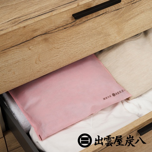 木質抽屜中放置了一個粉紅色的出雲屋炭八除濕木炭袋，袋身材質為細緻的不織布，袋上印有品牌名稱與調濕木炭字樣，周圍伴有白色與米色的衣物，整體呈現出整潔且具居家收納感的畫面。