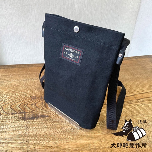 黑色帆布平板收納側背包，包包正面有一個標籤，上面印有日文圖案與文字，右下角有品牌標誌。