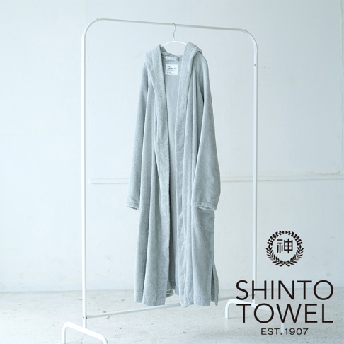 一件淺灰色連帽浴袍，採用蓬鬆有機棉材質，垂掛在白色衣架上，浴袍有兩個對稱的口袋，右側有品牌標誌。