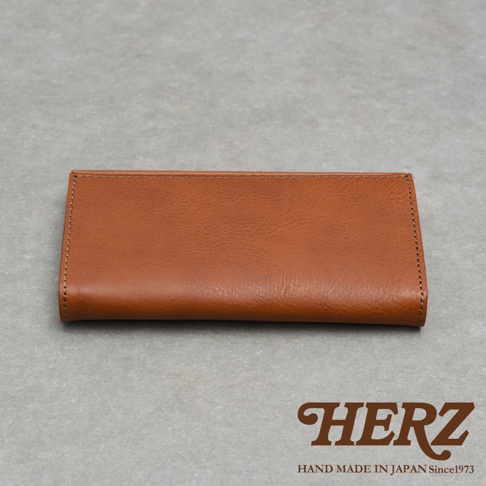 一張日本HERZ品牌職人手作皮革長夾的正面平拍圖，長夾呈現簡約長方形，選用溫潤的棕色皮革製作，表面有細緻的皮革紋理，邊緣處帶有規整的縫線細節，整體色調統一且質感厚實，右下方印有HERZ與手作日期標誌。