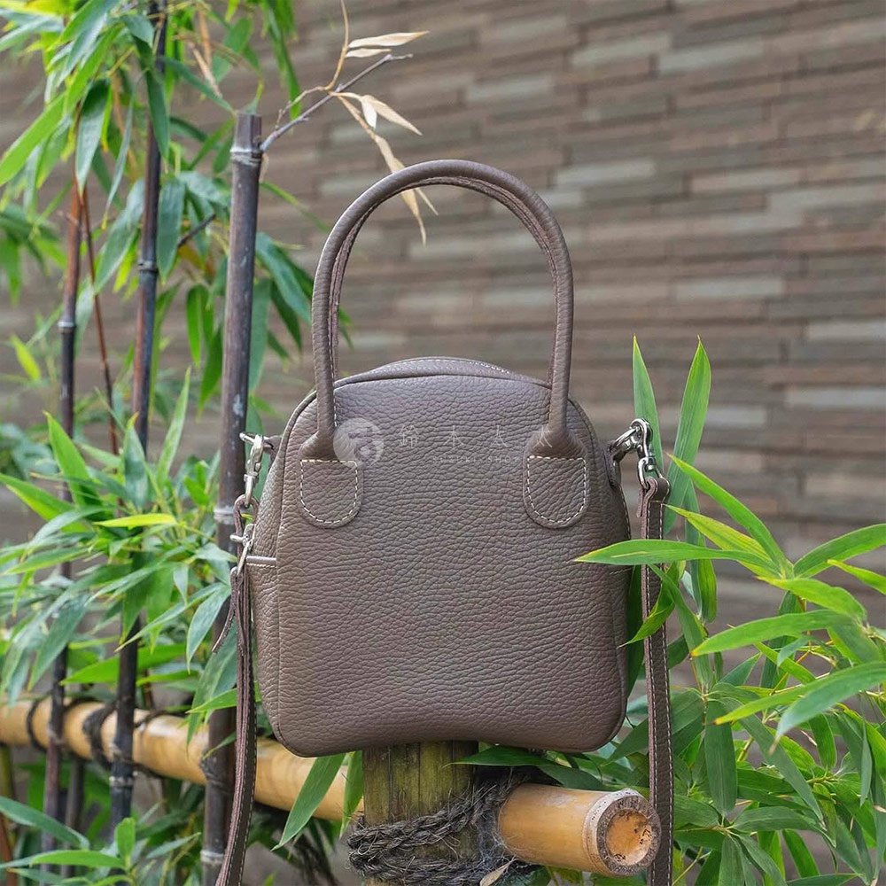 深棕色皮革製成的貝殼狀手提包，有兩個短提把和一條可拆卸長背帶，包身上有縫線裝飾。