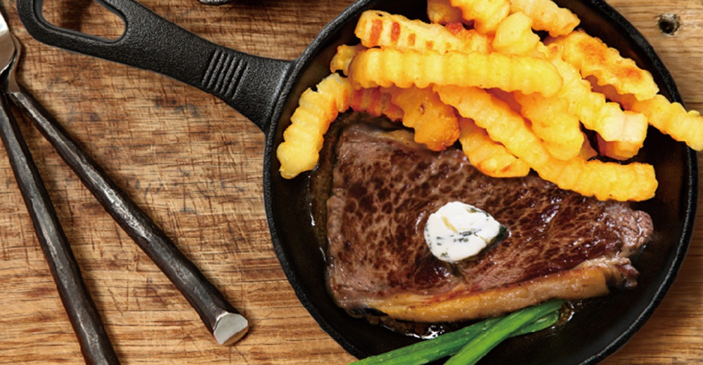 黑色鑄鐵平底鍋盛著一塊牛排，上面放著一塊奶油，旁邊是炸薯條和青豆。